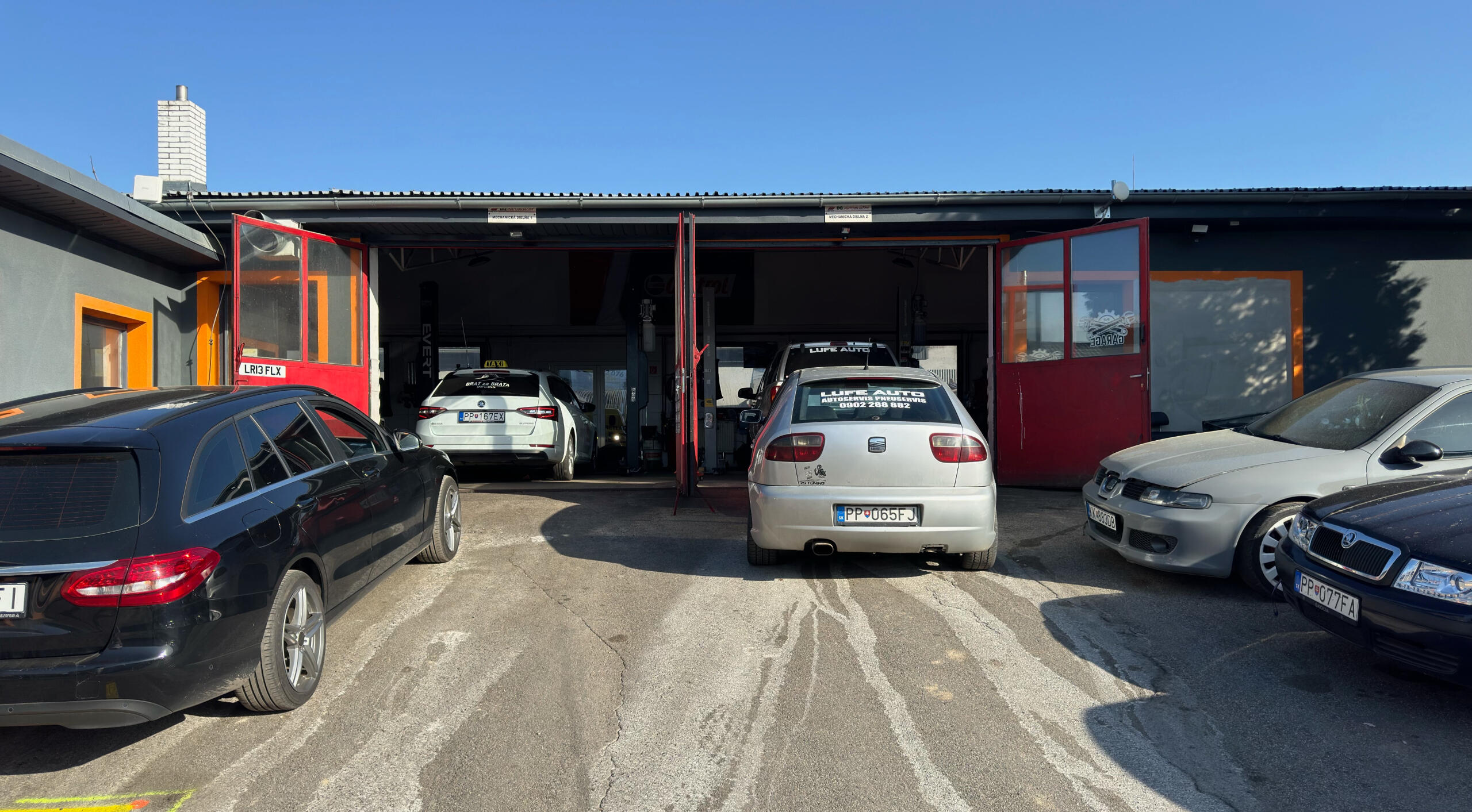 Skupina áut zaparkovaných pred garážou. Autá sú rôznych značiek a modelov. Niektoré autá sú nové a iné staršie. Garáž je priestranná. Pred garážou rastie strom.